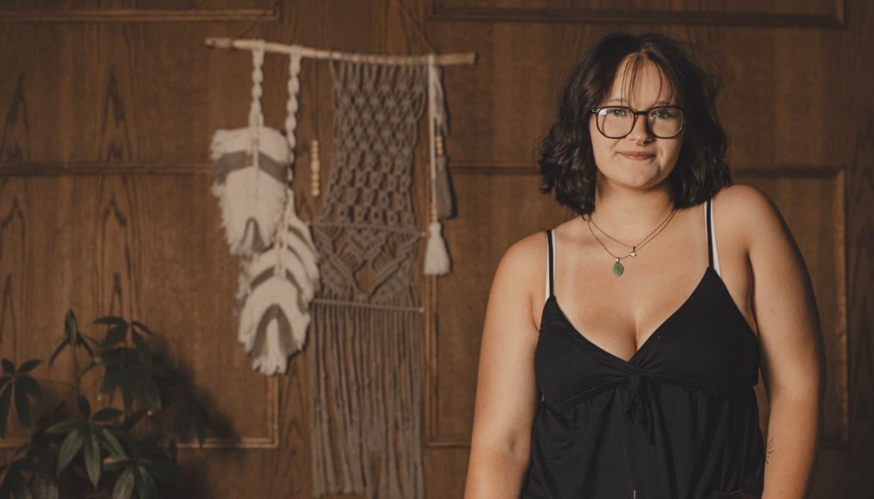 Smiling woman with glasses and dark hair stands before a wood-paneled wall with macramé decor.